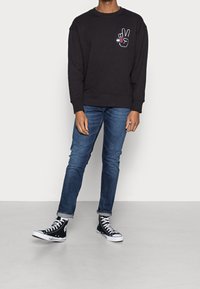 Persona che indossa una felpa nera con un logo di una mano della pace, jeans blu arrotolati e sneaker alte nere, in piedi su un pavimento grigio.