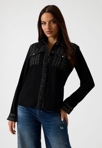 Zwarte shirt met contrasterend gestructureerd ruitpatroon op de schouders en zakken. Lange mouwen met gedetailleerde manchetten. Draagt bij blauwe distressed jeans.