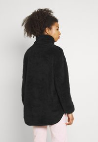 Pull noir et fluffy en matériau doux avec un col haut et des manches longues, légèrement plus long à l'arrière. Texture lisse, coupe décontractée.