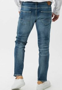 Jeans de denim azul de corte ajustado con detalles de desgastado, cinco bolsillos y una etiqueta de cuero marrón en la parte trasera de la cintura. Combinados con zapatillas blancas.