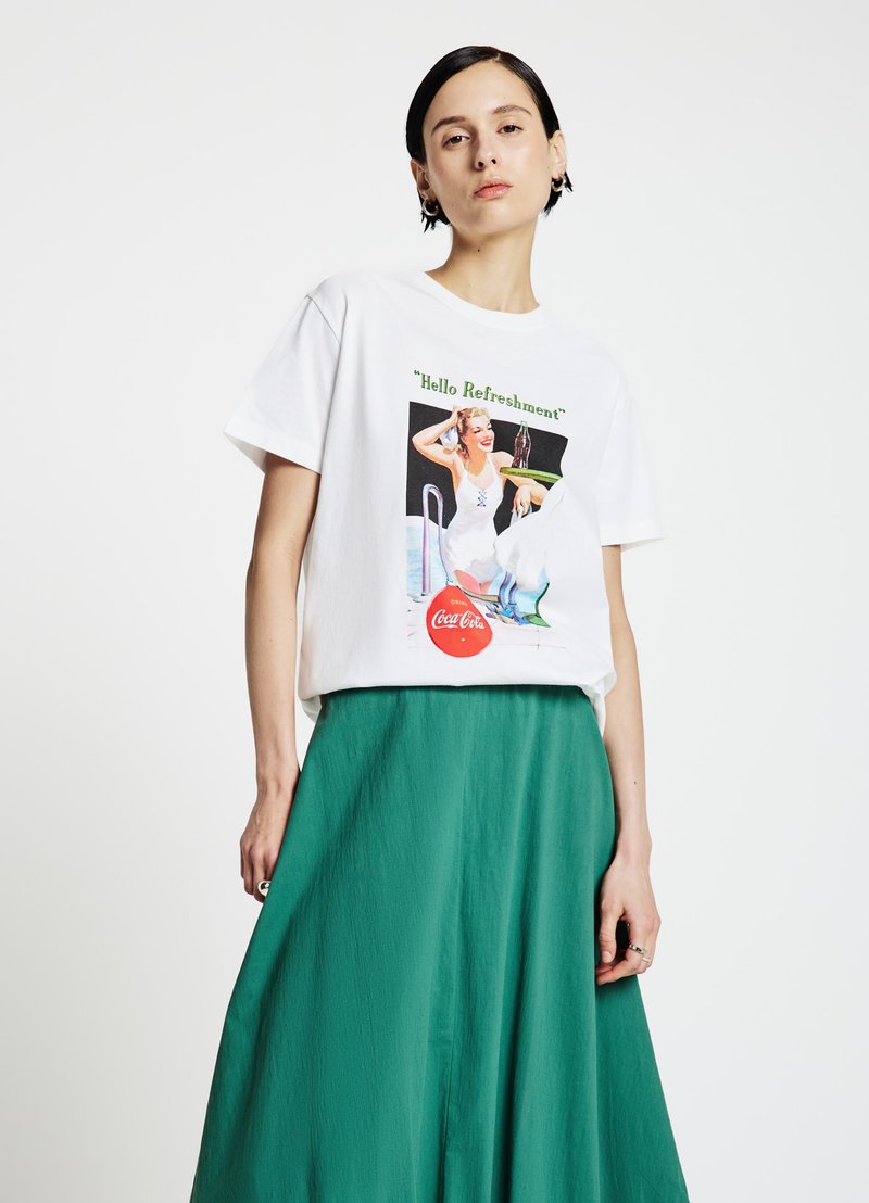 T-shirt en coton blanc arborant un graphisme rétro coloré avec le texte « Hello Refreshment » et un logo Coca-Cola, accompagné d'une jupe verte.