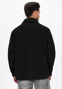 Mann, der mit dem Rücken zur Kamera steht, trägt eine schwarze Jacke mit Kragen und graue Jeans vor einem weißen Hintergrund.