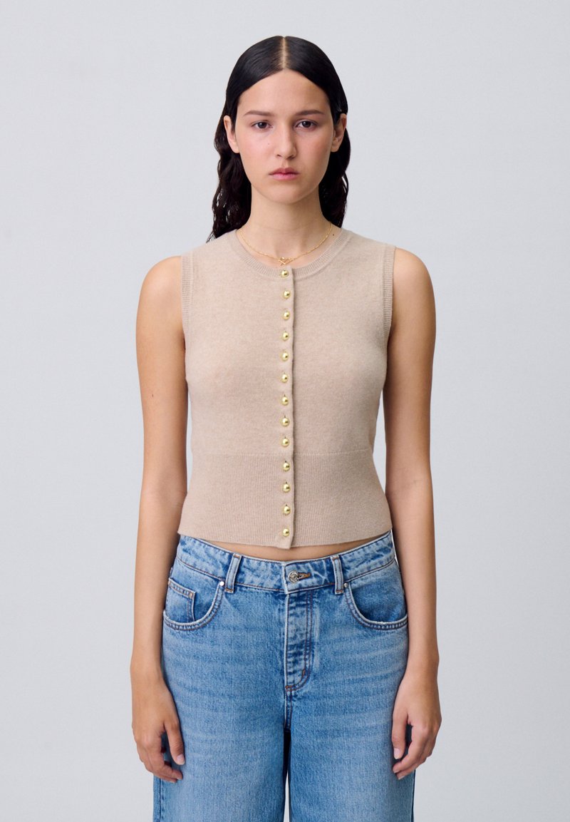 Top corto sin mangas en color beige, con escote redondo y cierre de botón dorado en la parte delantera, combinado con jeans de mezclilla azul claro.