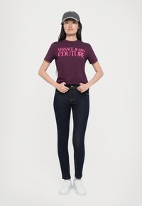 Violetinė marškinė su dideliu rožiniu tekstu "VERSACE JEANS COUTURE", derinami su tamsiai mėlynomis aptemptomis džinsais ir baltais sportbačiais. Modelis dėvi pilką kepurę.