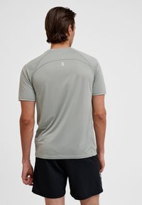 Hellgraues Sportshirt mit kurzen Ärmeln und rundem Ausschnitt, mit Raglan-Nähten und einem kleinen weißen Logo auf dem Rücken. Getragen mit schwarzen Shorts.