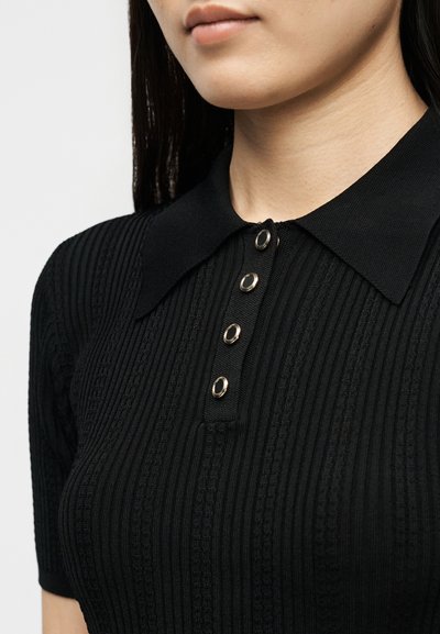 Κοντινό ενός ατόμου που φοράει μαύρο ριμπ πλεκτό πόλο με φαρδιά γιακά και τέσσερα ασημένια κουμπιά πατιλέτας.
