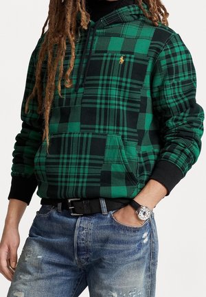 Osoba ubrana w zieloną i czarną kratowaną bluzę z kapturem z żółtym logo, niebieskie postarzane jeansy, czarny pasek oraz zegarek na rękę z czarnym paskiem.