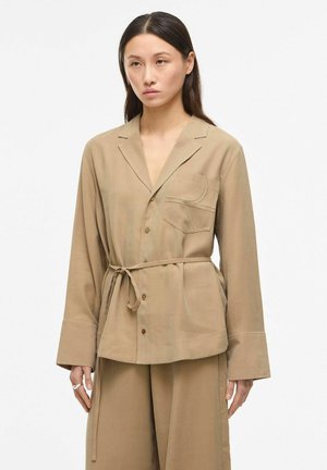 Kobieta w beżowej kurtce z paskiem, dużymi mankietami oraz pasujących szerokich spodniach na jednolitym białym tle.