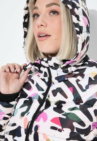 Veste matelassée avec un motif camouflage abstrait multicolore, dotée d'une fermeture éclair noire et d'une capuche réglable, fabriquée en matériau léger et brillant.