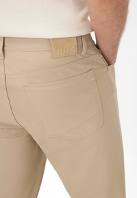 Beige katoenen broeken hebben een achterzak en een leren label met verhoogde merknaam. Naaddetails zijn zichtbaar op de achterkant en de zakken.