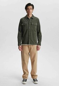 Jachetă cămașă din corduroy verde măslin, cu închidere cu nasturi și două buzunare la piept, asortată cu o pereche de pantaloni bej, lejeri, și adidași negri.
