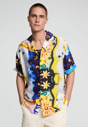 Junger Mann mit kurzen Haaren, der ein buntes kurzärmeliges Hemd mit Knöpfen trägt, mit Sonnen-, Mond- und Blumenmustern, die Hände in den Taschen.