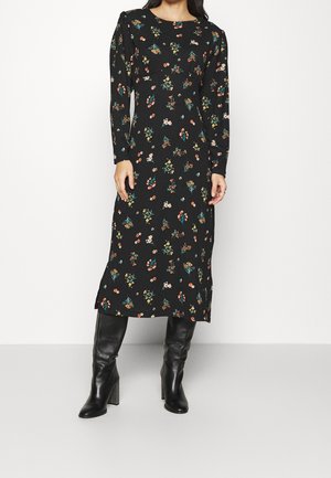 Kobieta w czarnej, midi sukience z długim rękawem w wielokolorowy kwiatowy wzór oraz w czarnych, wysokich kozakach na obcasie stoi na białym tle.