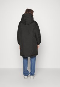 Černá puffer bunda s velkou kapucí, střední délka, hladká textura a elastické manžety, doplněná o světle modré džíny a tenisky.