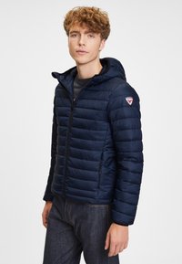 Ung man i en marinblå, vadderad huvtröja och mörka jeans, stående mot en ren vit bakgrund.