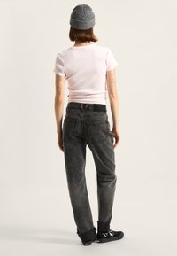 Camiseta de algodón de color rosa claro con canalé, jeans desgastados grises de corte holgado y zapatos negros. Gorra de lana gris, tatuajes visibles en los brazos.