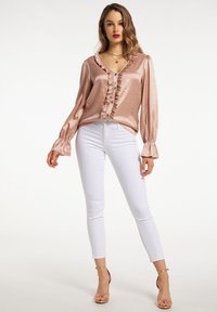 Blusa rosa oro arricciata con texture a motivo, maniche lunghe a palloncino e chiusura con bottoni, abbinata a pantaloni bianchi aderenti e tacchi color nude.