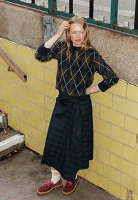 Pull en motif argyle marron et marine, jupe enveloppante à carreaux noirs, chaussures bordeaux avec des lacets roses, et chaussettes blanches avec un motif rouge.