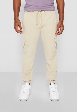 Mann trägt beige Cargohosen mit Kordelzug in der Taille und elastischen Bündchen, kombiniert mit weißen Turnschuhen und einem lila Hemd.
