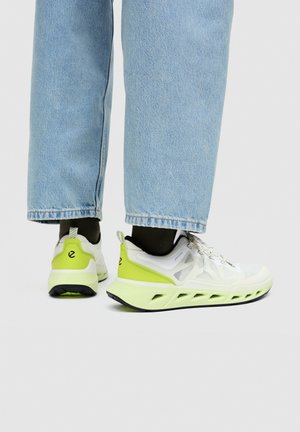 Person, die hellblaue Jeans und weiße Sneaker mit neon-grünen Akzenten trägt, steht vor einem schlichten hellgrauen Hintergrund.