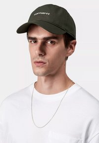 Gorra verde oliva con visera plana y logo blanco, combinada con una camiseta blanca sencilla y un collar de cadena de plata.