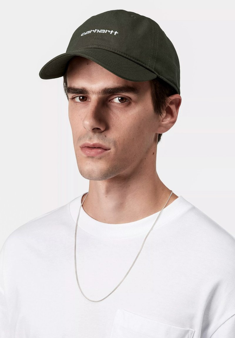 Gorra verde oliva con visera plana y logo blanco, combinada con una camiseta blanca sencilla y un collar de cadena de plata.