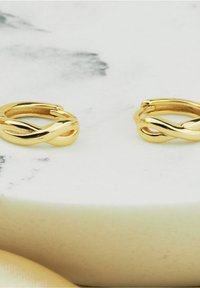 Guld vridna hängande örhängen med slät yta och sammanflätad design. Monterade på en ljus marmor yta.