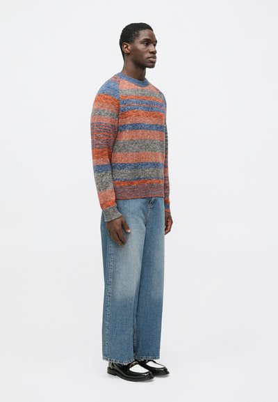 Pruhovaný pletený svetr v oranžové, šedé a modré barvě s žebrovanými manžetami a výstřihem. Kombinován s širokými světlemodrými džínami a černými botami.