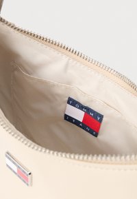 Bézs cipzáros táska belseje, sima anyagbélés látható, valamint egy kék címke piros és fehér részletekkel, rajta a "TOMMY JEANS" felirat.