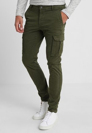 Mężczyzna noszący oliwkowozielone spodnie bojówki o dopasowanym kroju z klapkami na kieszeniach, do tego białe wysokie trampki i pasiastą koszulę z długim rękawem.