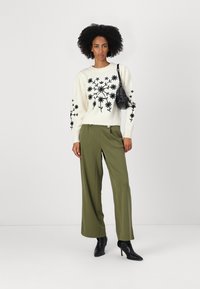 Maglione lavorato a maglia crema con ricami floreali neri, abbinato a pantaloni larghi verde oliva. La modella tiene una piccola borsa nera e indossa stivaletti neri.