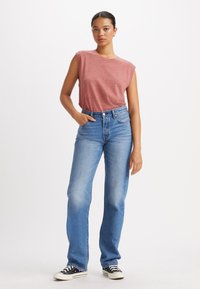 Rode, mouwloze t-shirt met ronde hals, gecombineerd met lichtblauwe recht gesneden jeans en zwart-witte lage sneakers. Eenvoudige, casual outfit.