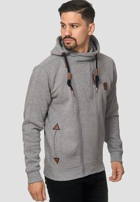 Hoodie zippé gris en tissu doux avec un col haut, présentant des accents en cuir marron et des patches triangulaires, ainsi qu'un cordon de serrage avec des embouts en métal.