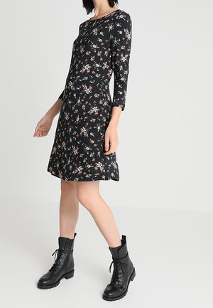 Frau in einem schwarzen Blumenkleid mit Dreiviertelärmeln und schwarzen geschnürten Kampfstiefeln, die vor einem schlichten hellen Hintergrund steht.