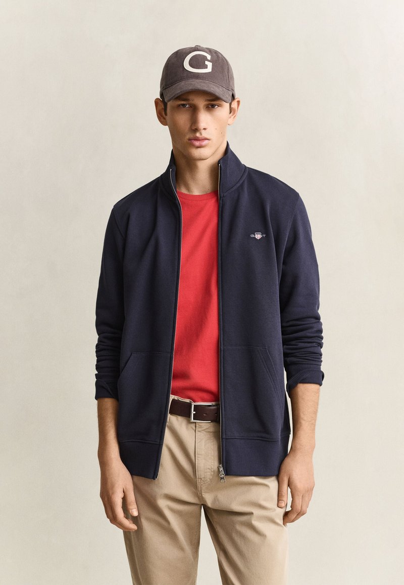 Junger Mann trägt eine dunkle Baseballkappe, eine marineblaue Jacke mit Reißverschluss, ein rotes Hemd, beigefarbene Hose und einen braunen Gürtel, steht vor einem schlichten hellen Hintergrund.