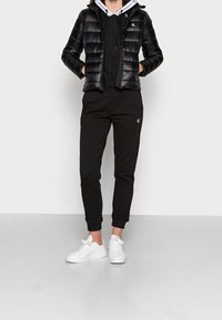 Svart puffjacka med quiltat mönster, svart sweatshirt under, svarta joggers och vita sneakers. Minimalistiska logotypdetaljer på kläderna.