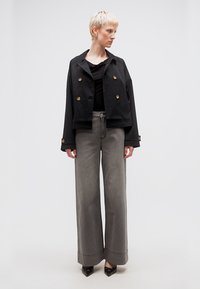 Άτομο με κοντά ξανθά μαλλιά που φοράει μαύρο κοντό σακάκι με διπλό πέτο, μαύρο τοπ, φαρδιά γκρι παντελόνια και μαύρα μυτερά παπούτσια, στέκεται και κοιτάζει δεξιά.