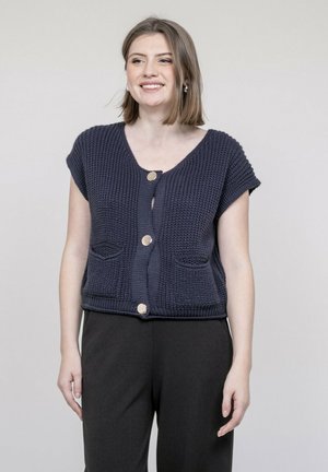 Femme souriante, portant un cardigan en maille bleu marine à manches courtes avec de gros boutons et un pantalon noir, debout devant un fond uni.