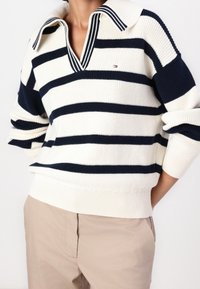 Henkilö, jolla on päällä vaalean kermanvärinen ja tummansininen raidallinen neulepaita, jossa on kaulus, sekä vaaleat housut. Kädet taskuissa taustalla on yksinkertainen tausta.