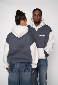 Hanorac supradimensionat gri închis și alb, cu o cusătură în formă de V. Asortat cu blugi albaștri care au accente brodate pe buzunare.