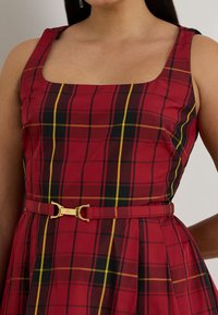 Sarkans rūtainas kleita ar kvadrātveida izgriezumu, ar dzeltenām un melnām līnijām. Plāna josta ar zelta metāla detaļām akcentē vidukli. Kokvilnas audums.