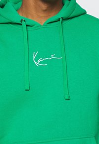 Sudadera verde hecha de una tela con textura suave. Presenta una capucha con cordón y un logo bordado en blanco en el pecho.