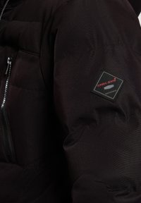 Czarna kurtka puchowa z pikowanymi sekcjami, z zamkiem z przodu i kwadratową naszywką z logo na lewym rękawie. Materiał jest gładki i lekko błyszczący.