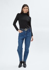 Schwarzes langärmliges Rollkragenoberteil, blaue hochgeschnittene Jeans mit Used-Look, silberner Verzierte Gürtel, kombiniert mit schwarzen Ankle Boots.