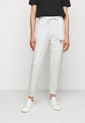 Osoba nosząca białe spodnie dresowe z regulowanym sznurkiem, kieszeniami po bokach i logo na udzie, w połączeniu z białymi trampkami i czarnym krótkim topem.