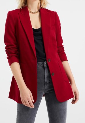 Person wearing a red blazer over a black top and gray high-waisted jeans, accessorized with a gold chain necklace.
