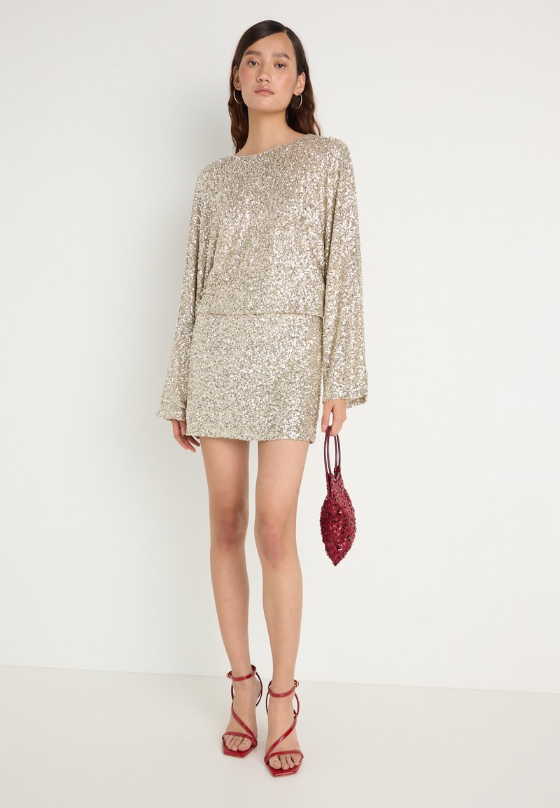 Svetleča zlata obleka iz bleščic z dolgimi rokavi in raven kroj, v kombinaciji z teksturirano bordo ročno torbo in rdečimi čevlji na peto.