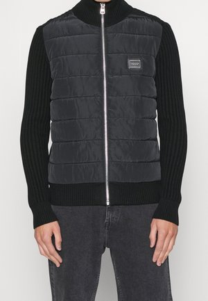 Mann trägt eine schwarze gesteppte Zip-up-Weste über einem schwarzen gerippten Langarmpullover, kombiniert mit dunkelgrauen Jeans.