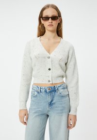 Kerman ribbattu cardigan cu uânn mu keep maig ra dhip V, chhe nai buttons, e chhe cropped fit. Arhivêz chhe ek khawôḍī bālan sari with subtle speckled accents.