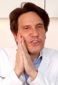 Mannlig helsepersonell i hvit frakk, hender på ansiktet, med kort mørkebrunt hår og skjorte med blå striper. Myk bakgrunnsbelysning.